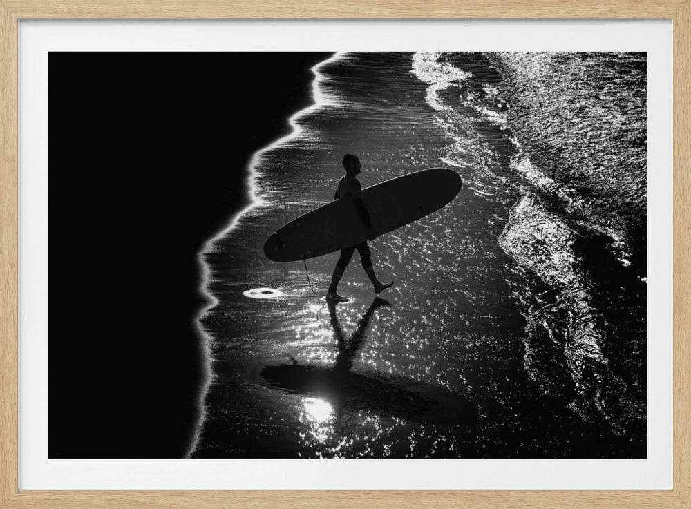 A dramatic, high-contrast, black and white aerial view of a silhouetted surfer carrying their surfboard along the shoreline. The sun glints off the wet sand and the dark water, creating a sparkling effect and a clear reflection of the surfer. Poster