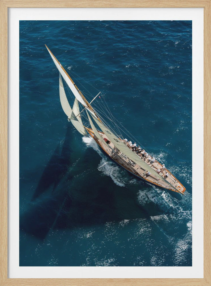 An aerial, high-angle photograph of a classic wooden racing yacht sailing on a vibrant blue ocean. The boat is heeled over, cutting through the water and creating a white wake, with its large sails full of wind and numerous crew members visible on deck. Decor