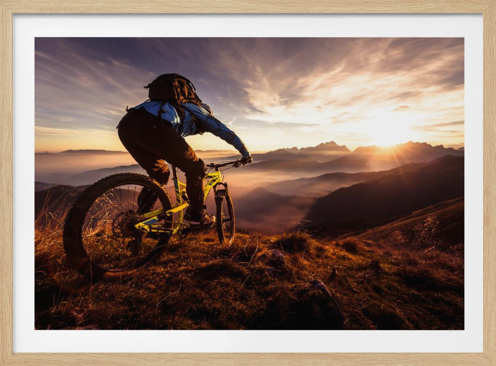 A mountain biker, seen from behind, stops at the top of a grassy hill to admire a spectacular sunset over a vast mountain range. The valleys below are filled with mist, and the sun's golden rays create a stunning starburst effect against the silhouetted peaks. Print