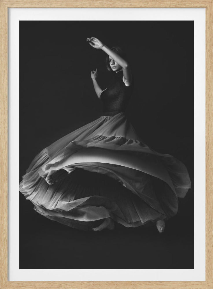 A dramatic black and white photograph of a woman dancing in a studio setting. She wears a dark, fitted top and a long, flowing light-colored skirt that swirls around her as she moves, creating beautiful folds and shadows. Her arms are raised in a graceful pose as she looks towards the camera against a solid dark background. Print
