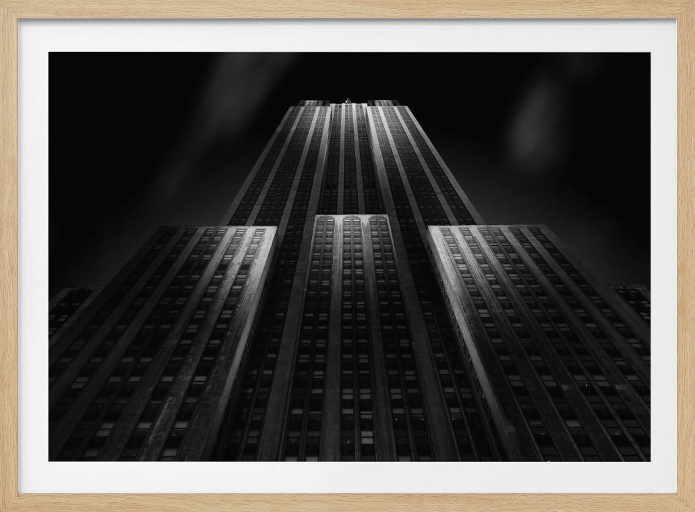 A dramatic, low-angle black and white photograph of the Empire State Building at night. The perspective looks up from the base, emphasizing the skyscraper's height and Art Deco details. Spotlights highlight the vertical lines of the building against a dark, moody sky, all enclosed in a silver frame. Poster