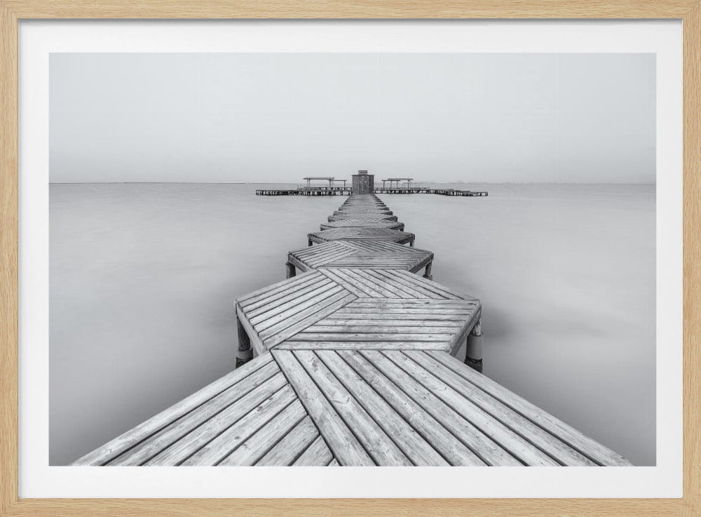 A framed, black and white photograph capturing a long wooden pier from a first-person perspective. The pier, constructed from a series of hexagonal platforms, stretches out into a calm, placid body of water that disappears into a foggy horizon, creating a serene and minimalist scene. Print