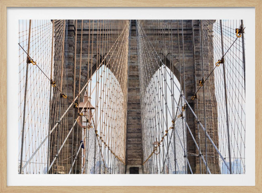 A framed, close-up photograph of the Brooklyn Bridge, focusing on the symmetrical view through the web of suspension cables towards the iconic stone Gothic arches of the towers. Print