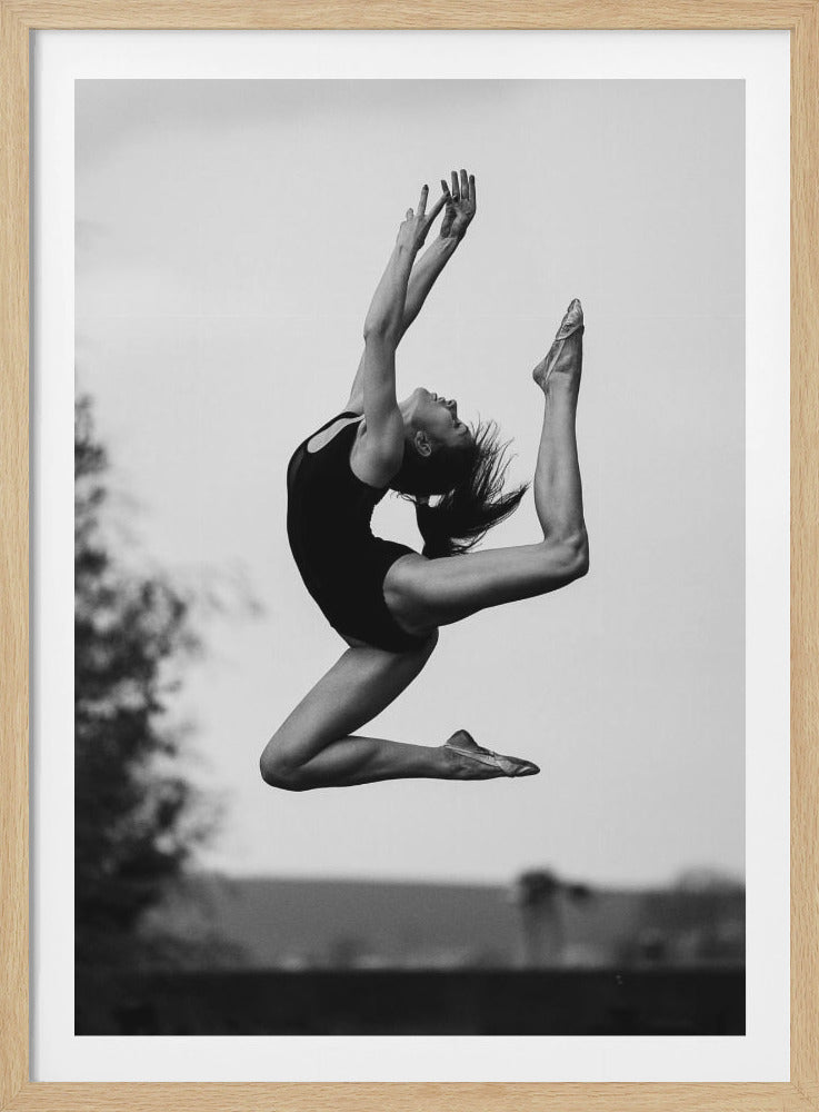A black and white photograph of a graceful ballerina mid-leap, with her body arched back, arms raised, and one leg extended high. The dancer is wearing a dark leotard and the image is framed in black. Wall Art