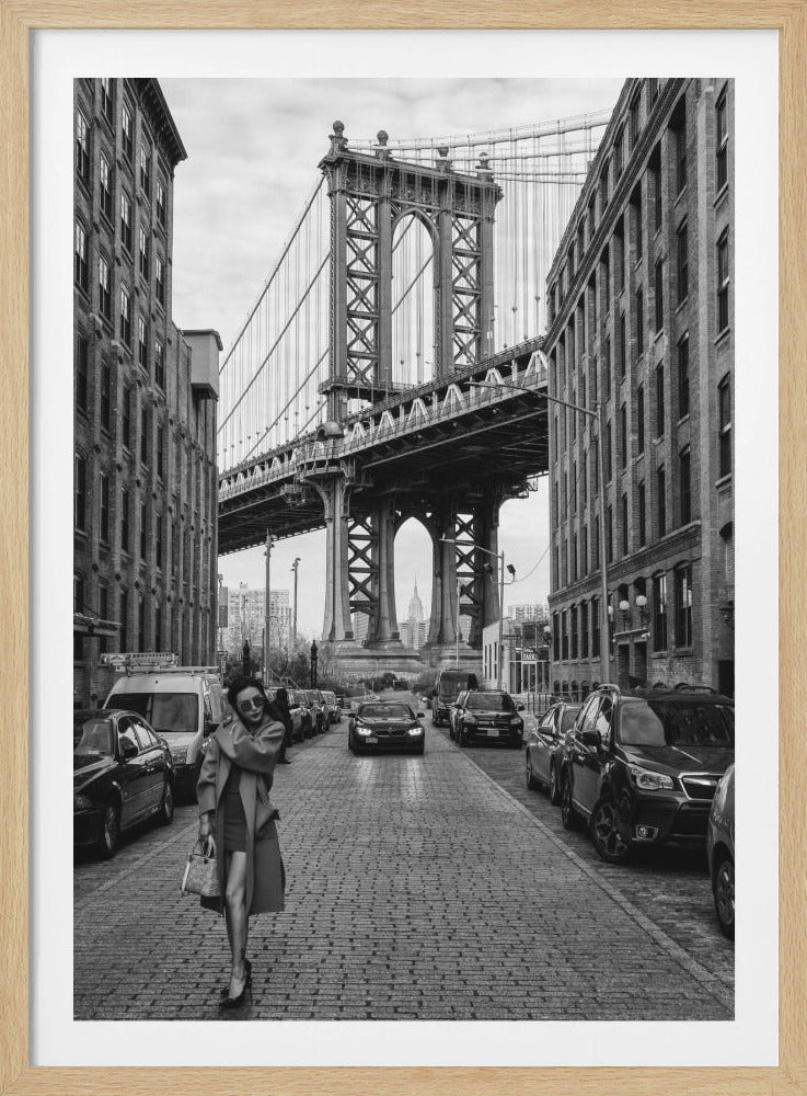 A black and white vertical photograph of a stylish woman walking down a cobblestone street in DUMBO, Brooklyn. The massive Manhattan Bridge looms in the background, framed by tall brick buildings, with the Empire State Building visible through one of its arches. Print