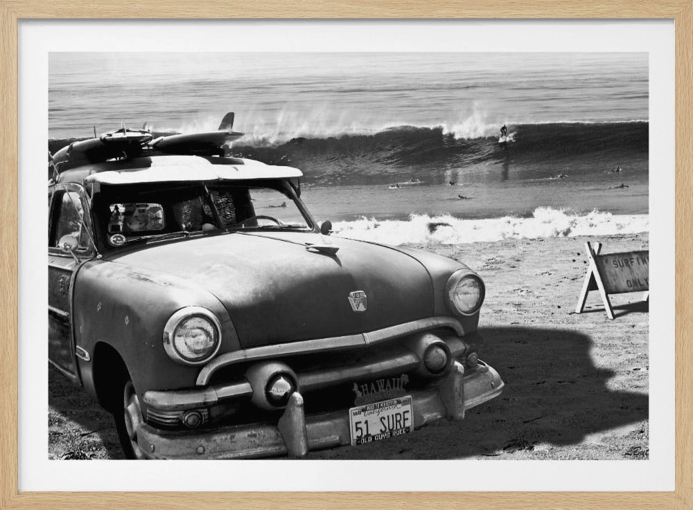 A vintage black and white photograph of a classic car parked on a sandy beach with surfboards strapped to the roof. In the background, a large wave is breaking with a lone surfer riding it, while other surfers are in the water. A sign on the beach reads 'SURFING ONLY'. Decor