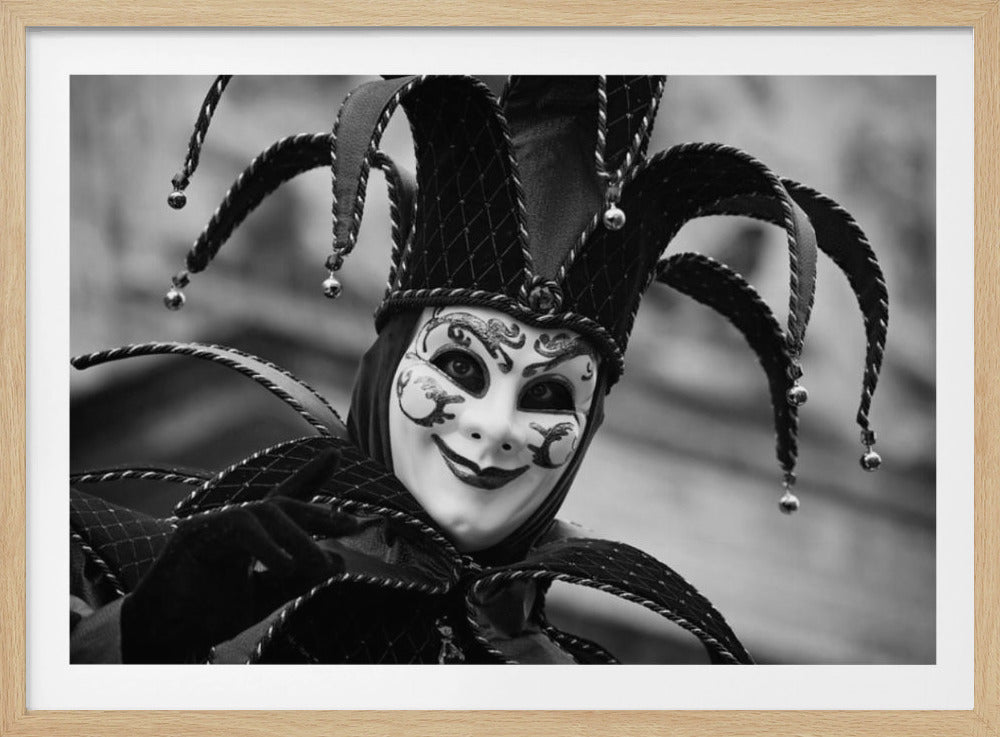 A close-up, black and white photograph of a person wearing an elaborate Venetian jester costume and mask. The white mask features intricate black scrollwork, a mischievous smile, and dark, expressive eyes looking directly at the viewer. The costume includes a classic jester hat with multiple points adorned with small bells, and the figure's black-gloved hand is partially raised in a playful gesture. The entire image is presented within a silver, brushed-metal frame. Print