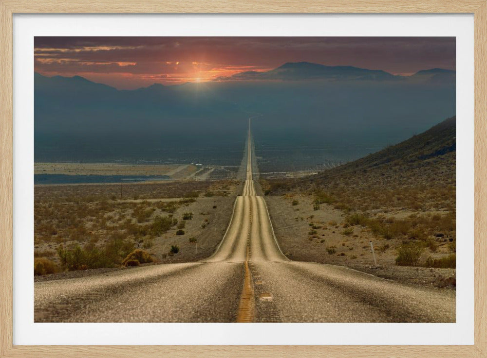 A high-angle view of a long, straight, and hilly road disappearing into the horizon of a vast desert valley at sunset, with mountains in the background under a warm, glowing sky, all enclosed in a silver frame. Poster