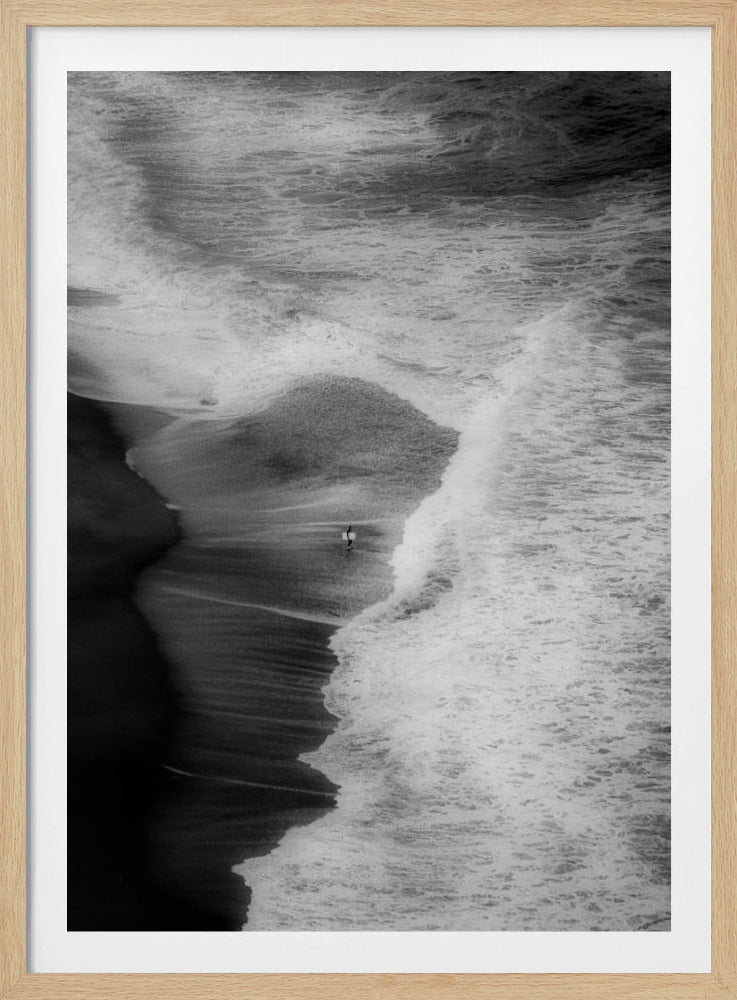 A high-angle, black and white photograph of a lone surfer carrying a surfboard at the water's edge on a dark sand beach. The vast, textured ocean with large, foamy waves crashing ashore dwarfs the solitary figure, creating a sense of scale and solitude. Decor