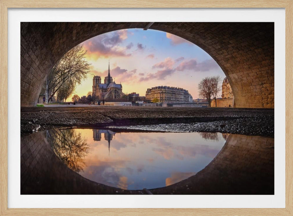 A unique view of Notre Dame Cathedral in Paris, framed by the arch of a stone bridge at sunset. In the foreground, a puddle on the ground perfectly reflects the iconic cathedral and the warm, colorful sky. Decor