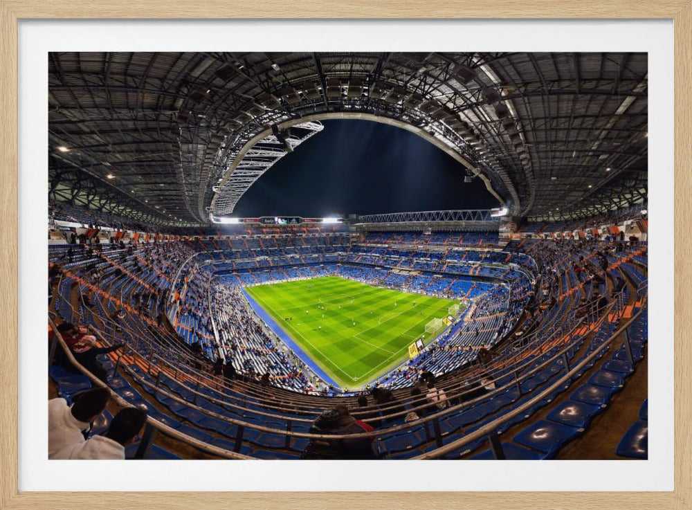 A sweeping, wide-angle photograph from the upper stands of a crowded soccer stadium at night, with the brilliant green pitch illuminated below and the grand architectural roof curving against the dark sky. The image is presented within a silver frame. Print