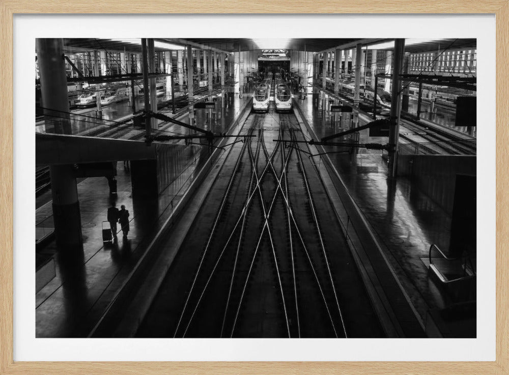 A high-angle, black and white photograph of a large, modern train station. Symmetrical train tracks lead away from the viewer towards two parked trains in the distance, with their reflections visible on the polished floor. To the left, a silhouetted couple walks along a platform with luggage. Artwork