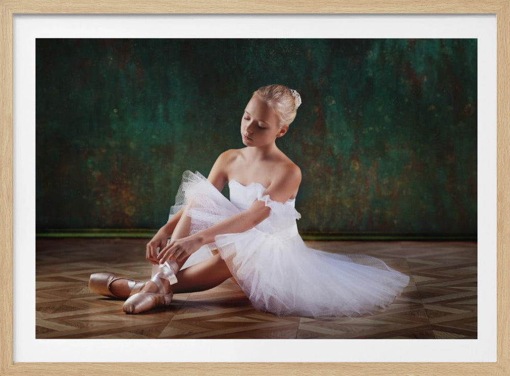 A framed photograph of a young blonde ballerina in a white tutu sitting on a polished wood floor. She is looking down while tying the ribbon on her pink pointe shoe against a dark, textured green background. Poster