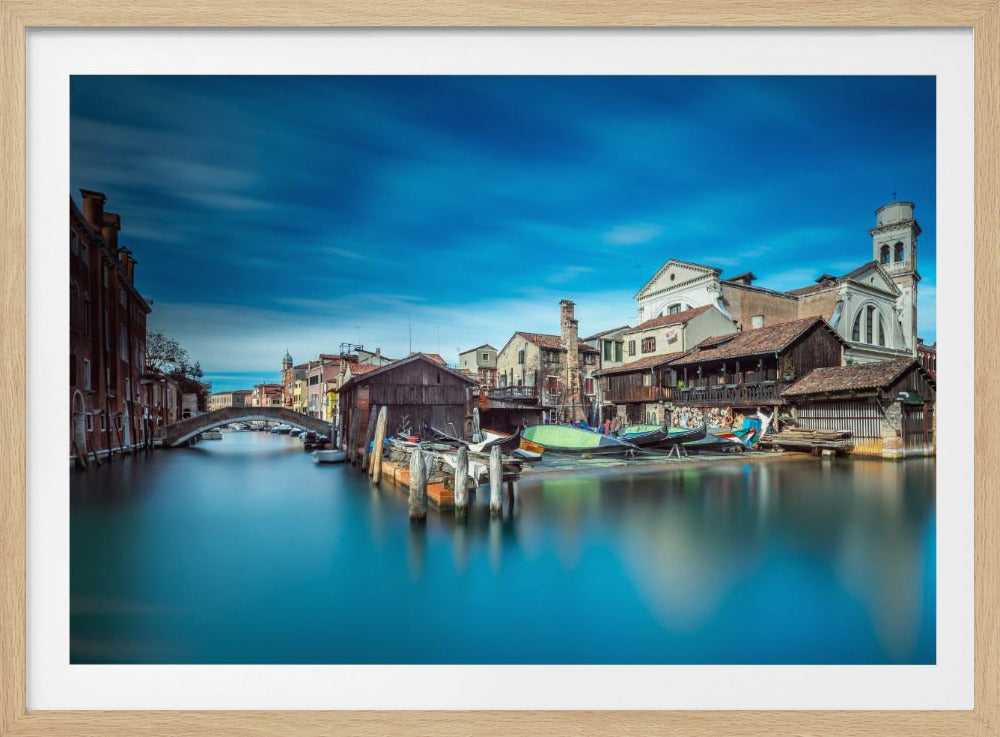 A long-exposure photograph of a tranquil Venetian canal, with silky smooth blue water reflecting rustic buildings, moored gondolas, and a distant stone bridge, all under a vibrant, streaky blue sky. Print