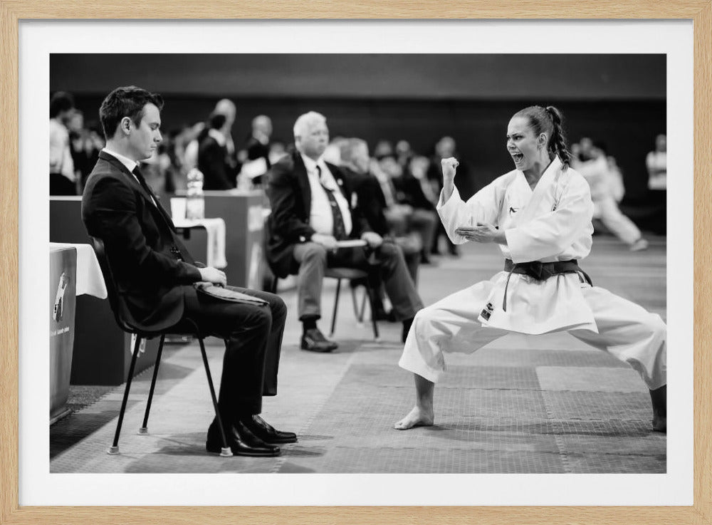 A dynamic black and white photograph of a female martial artist in a white gi and black belt, mid-kiai. She is in a deep, wide stance on a matted floor, her face filled with intensity and her fist clenched. In the foreground, a man in a suit sits and watches, while other spectators are seated in the blurred background, suggesting a competition or demonstration setting. The entire image has a silver-colored frame. Print