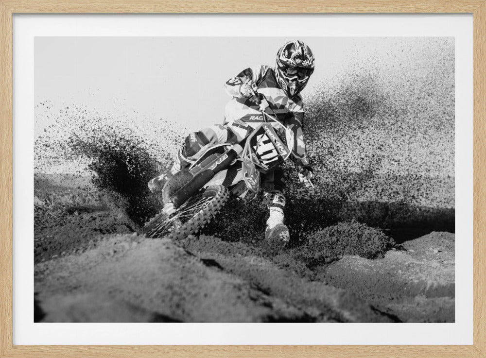 A dynamic black and white action photo of a motocross rider in full gear leaning hard into a turn, kicking up a large spray of dirt and sand from the track. The image is presented within a silver frame. Decor