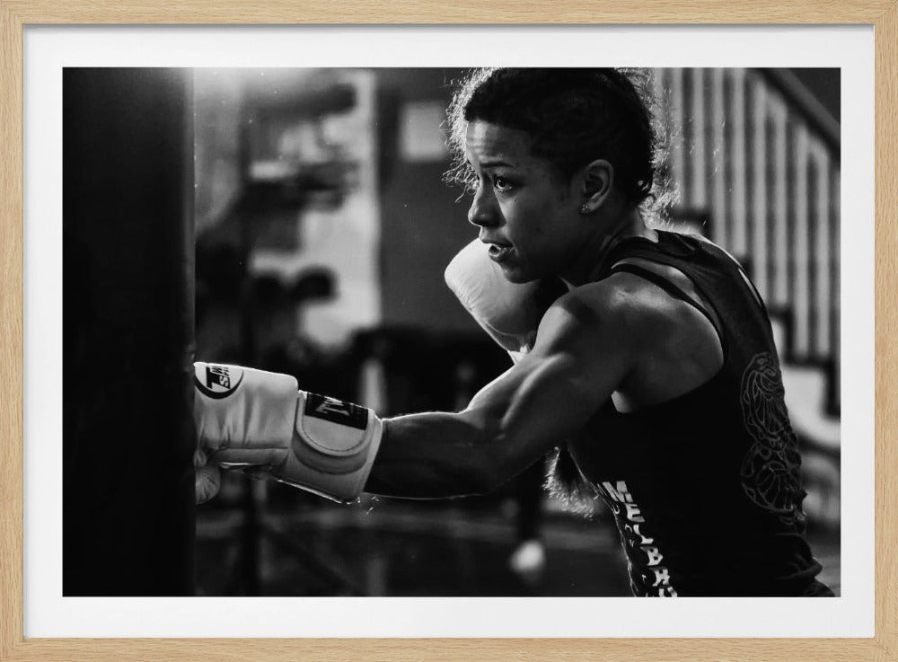 A dramatic black and white photo of a muscular female boxer in profile, intensely focused as she punches a heavy bag, set within a silver frame. Print