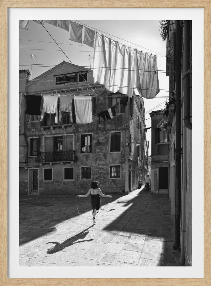 A black and white photograph capturing a girl from behind as she jump-ropes down a sunlit, narrow cobblestone street in Venice. Her long shadow is cast before her, and above, laundry hangs on clotheslines strung between old, weathered European buildings. Wall Art