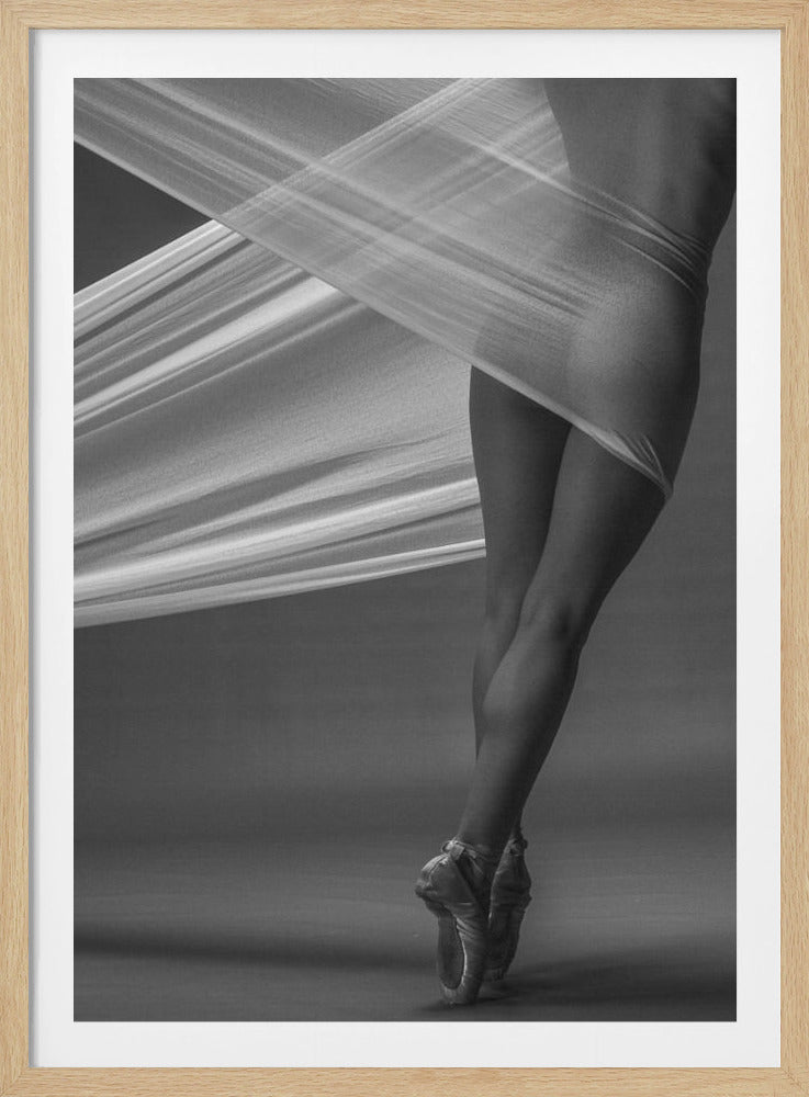 A dramatic black and white photograph of a ballerina's legs and torso. She is standing on pointe in ballet shoes, and her body is partially wrapped in a wide, sheer, white fabric that stretches across the image diagonally, creating a sense of movement and abstraction against a solid grey background. Artwork
