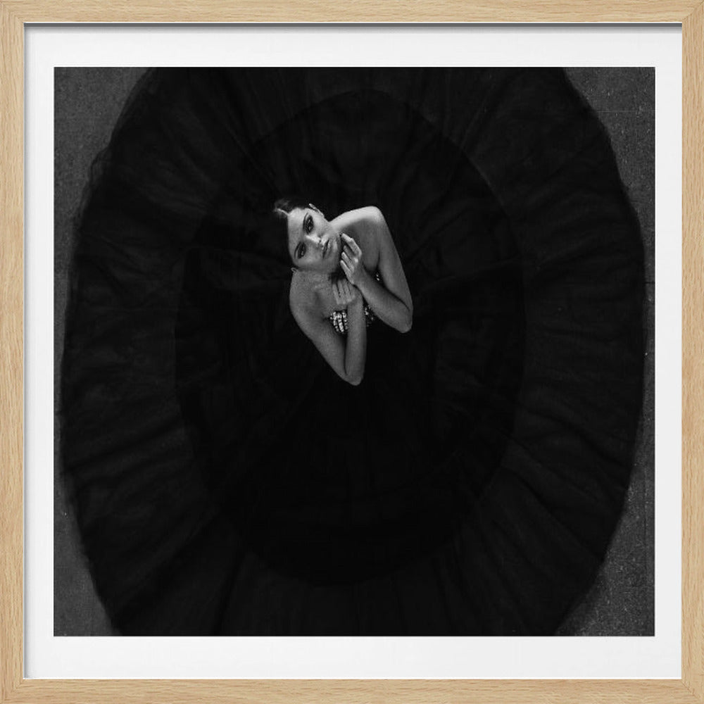 A striking black and white, bird's-eye-view photograph of a woman centered in a voluminous black gown. Her dress spreads out in a perfect circle, filling the dark, textured background. She looks directly up at the camera with an intense gaze. The square image is encased in a light wood frame. Wall Art