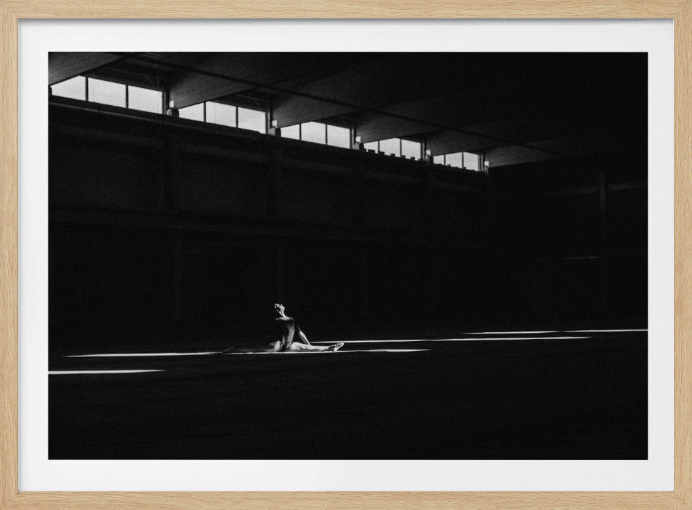 A dramatic black and white photograph of a lone gymnast stretching in a split pose on the floor of a large, dark gymnasium. Strips of bright light cut across the darkness, highlighting the athlete's form and the vast, empty space around them. Artwork