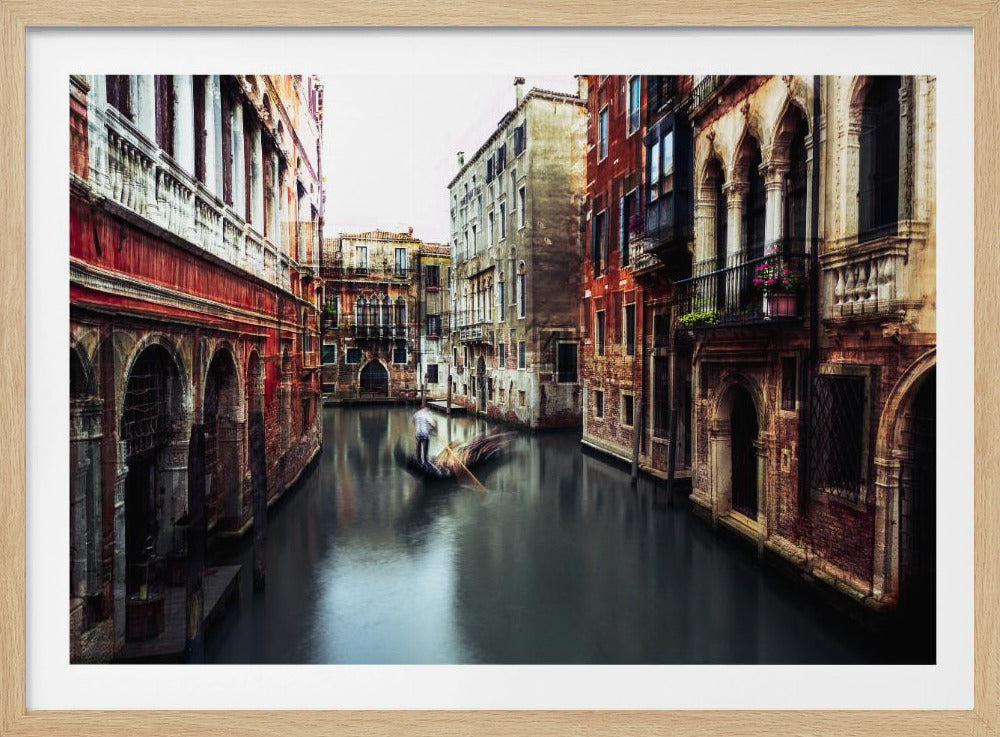 A long-exposure photograph of a gondolier steering a gondola through a narrow, dark canal in Venice, Italy. The motion creates a blur, contrasting with the sharp details of the historic, colorful buildings lining the waterway, all framed in a silver border. Decor