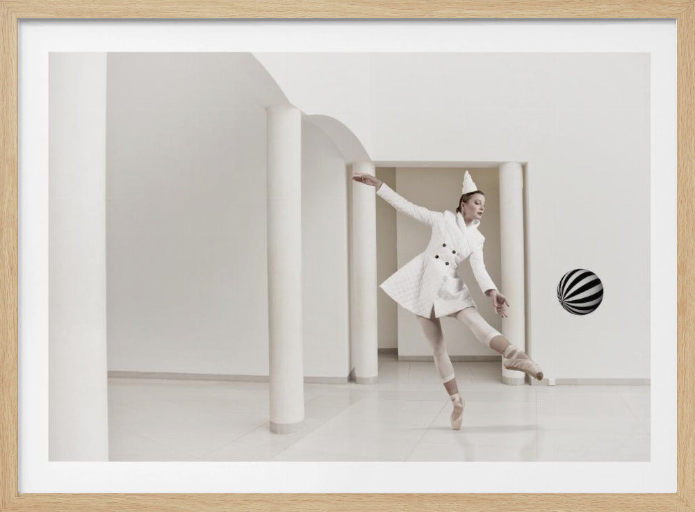 A ballerina dressed in a white quilted coat and a pointed hat dances en pointe in a minimalist white hall with large columns. She gracefully extends her arms, looking towards a black and white striped ball that appears to be floating in mid-air. The scene is presented within a silver picture frame. Wall Art