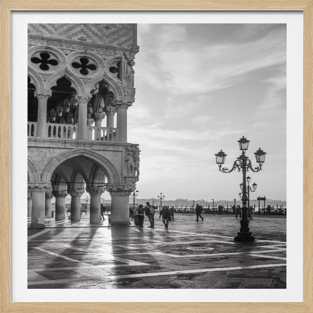 A framed black and white photograph of the Doge's Palace in Venice. The ornate gothic architecture is on the left, casting long shadows across a wet, reflective piazza. An old-fashioned lamppost stands on the right, with people strolling in the background near the water under a soft, cloudy sky. Artwork
