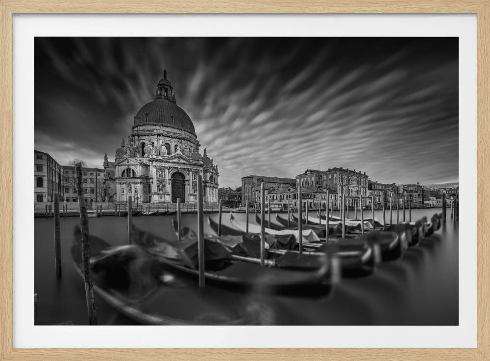 A dramatic black and white long-exposure photograph of Venice, featuring the Basilica di Santa Maria della Salute across the Grand Canal. In the foreground, a row of moored gondolas is partially blurred, leading the eye towards the historic buildings. The sky is filled with streaky, motion-blurred clouds, creating a sense of movement above the still water. The entire image is enclosed in a silver, textured frame. Wall Art