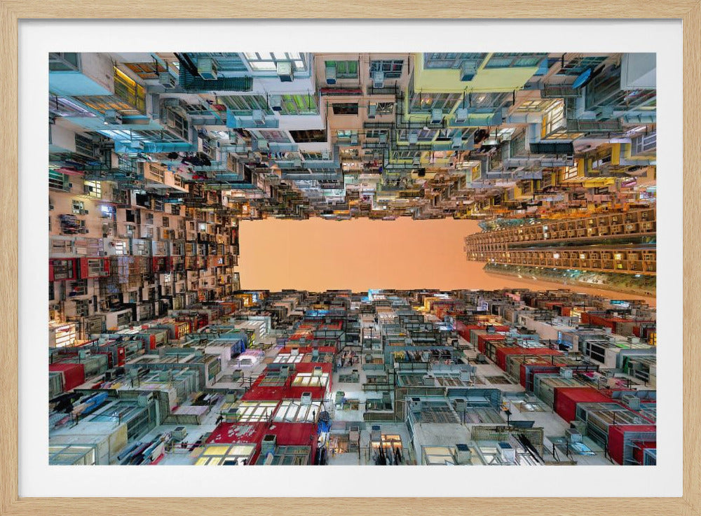 An upward-looking photograph from a courtyard surrounded by densely packed, multi-colored apartment buildings that create a rectangular frame around an orange sky. The unique perspective creates a surreal and geometric view of an urban landscape, all enclosed in a silver frame. Artwork