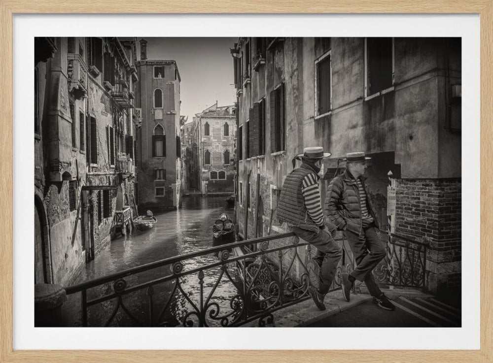 A framed, black and white photograph of Venice, featuring two gondoliers in traditional hats and striped shirts resting on an ornate bridge overlooking a narrow canal, with weathered historic buildings rising on either side. Print