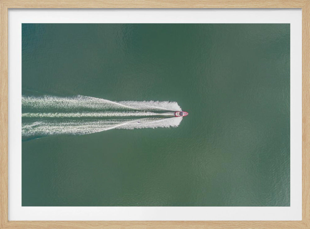 An aerial top-down photograph of a red motorboat speeding across a vast expanse of calm, green water, leaving a long, white, frothy wake behind it. The scene is presented within a simple silver frame. Print