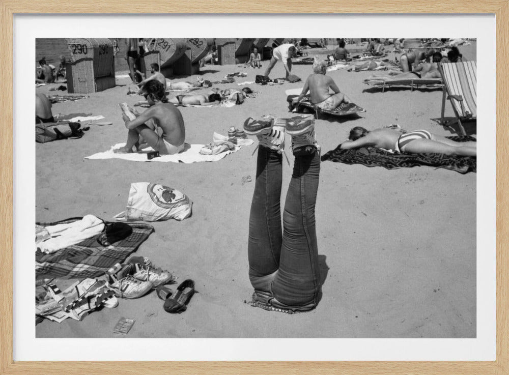 A quirky, vintage black and white photograph of a crowded beach. The central focus is a person lying on their back with their legs straight up in the air, oddly wearing jeans and sneakers. Surrounding them are numerous other beachgoers sunbathing on towels and in wicker beach chairs, creating a busy and candid summer scene. Wall Art