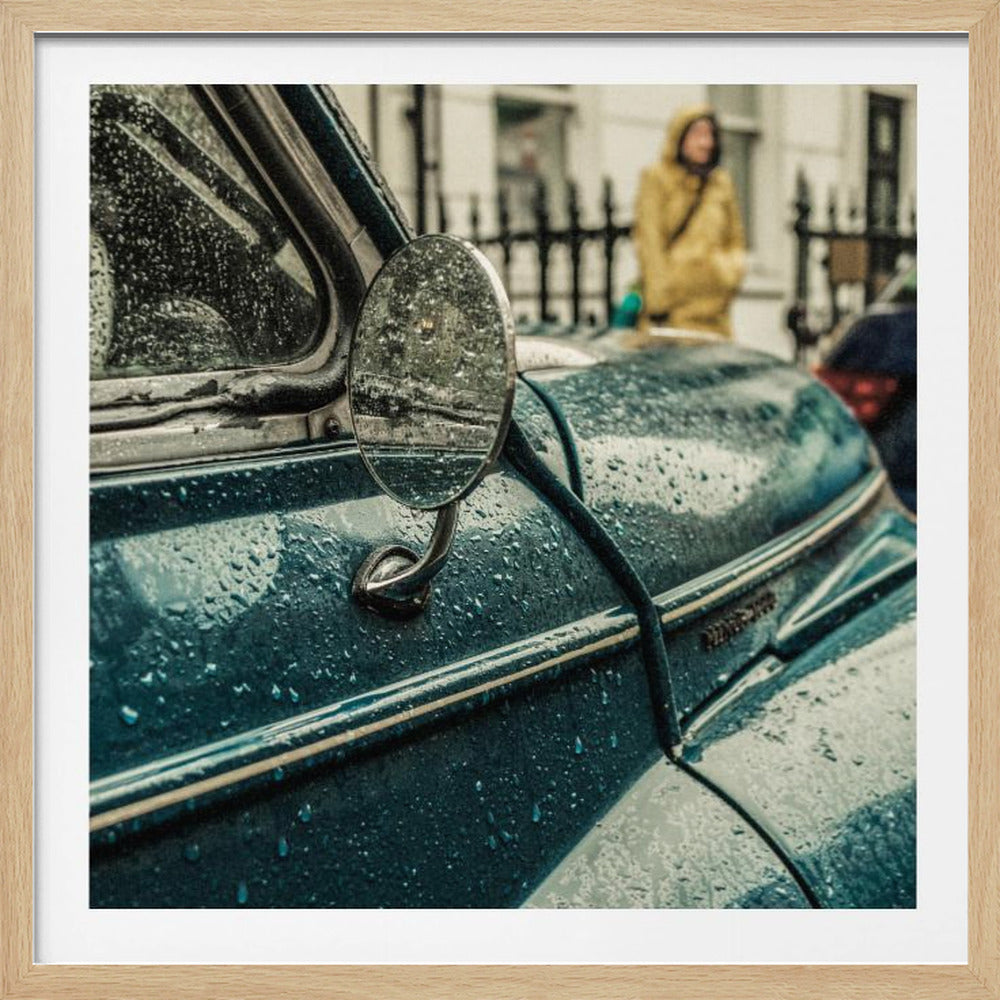 A close-up, moody photograph of a vintage teal car on a rainy day, with raindrops covering its surface. The oval side-view mirror reflects the wet street, and in the background, a person in a yellow raincoat is visible. Wall Art