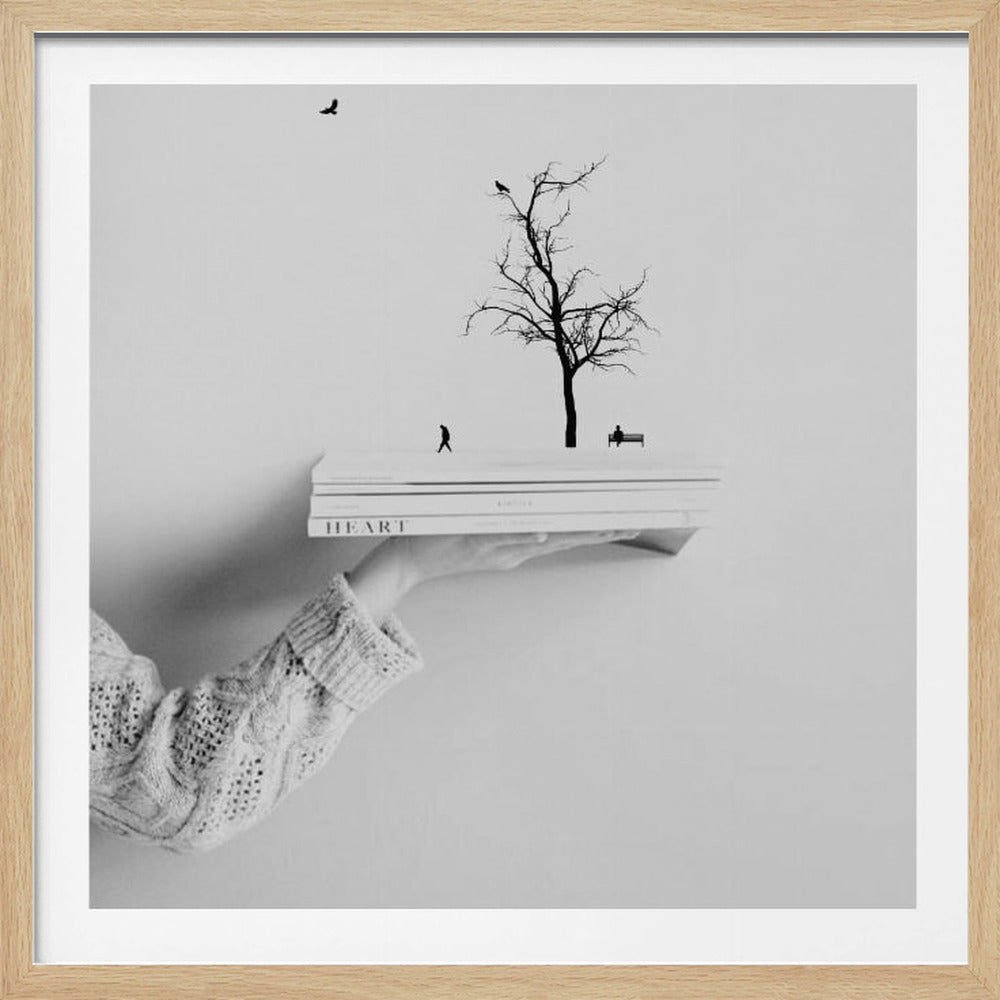 A framed, black and white conceptual art piece showing a hand in a knit sweater holding a stack of books. On top of the books is a miniature silhouette scene of a leafless tree, a person walking, another sitting on a bench, and birds flying above. Artwork