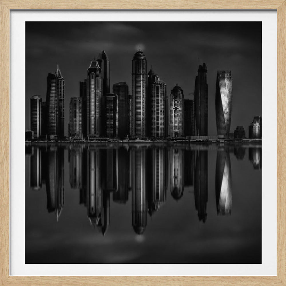 A dramatic black and white photograph of a modern city skyline at night, its towering skyscrapers perfectly reflected in the calm water below, all enclosed within a light brown wooden frame. Print