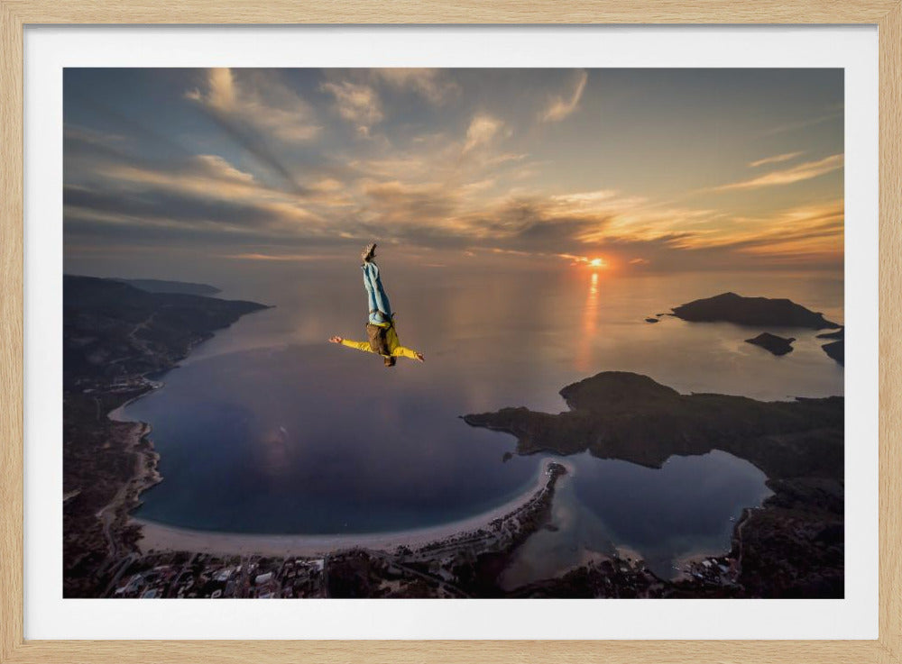 A breathtaking aerial view of a skydiver in a head-down position, freefalling over a vast coastal bay at sunset. The setting sun casts a golden glow over the ocean and clouds, highlighting the islands and coastline below. Decor
