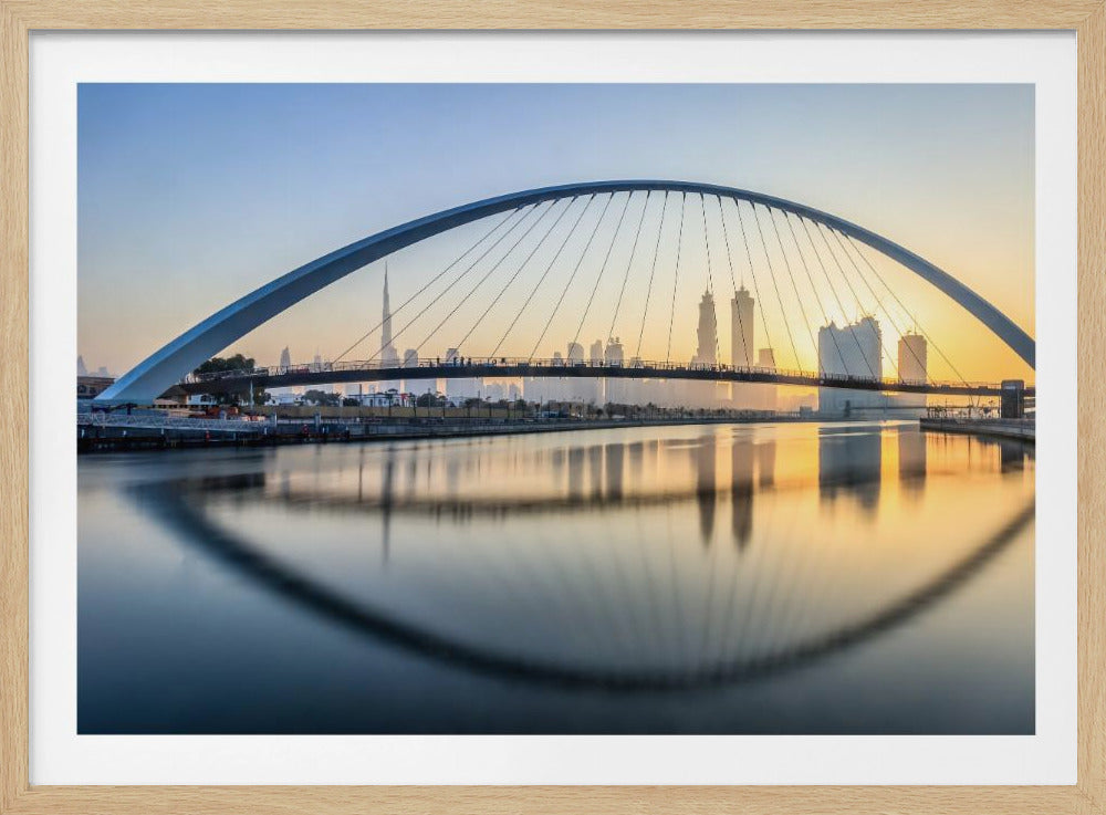 A framed photograph of the Dubai Water Canal Bridge at sunrise, with its large arch spanning across the water. The Dubai skyline, including the Burj Khalifa, is visible in the background, silhouetted against a golden and blue sky. The bridge and buildings create a perfect reflection on the calm surface of the water. Wall Art