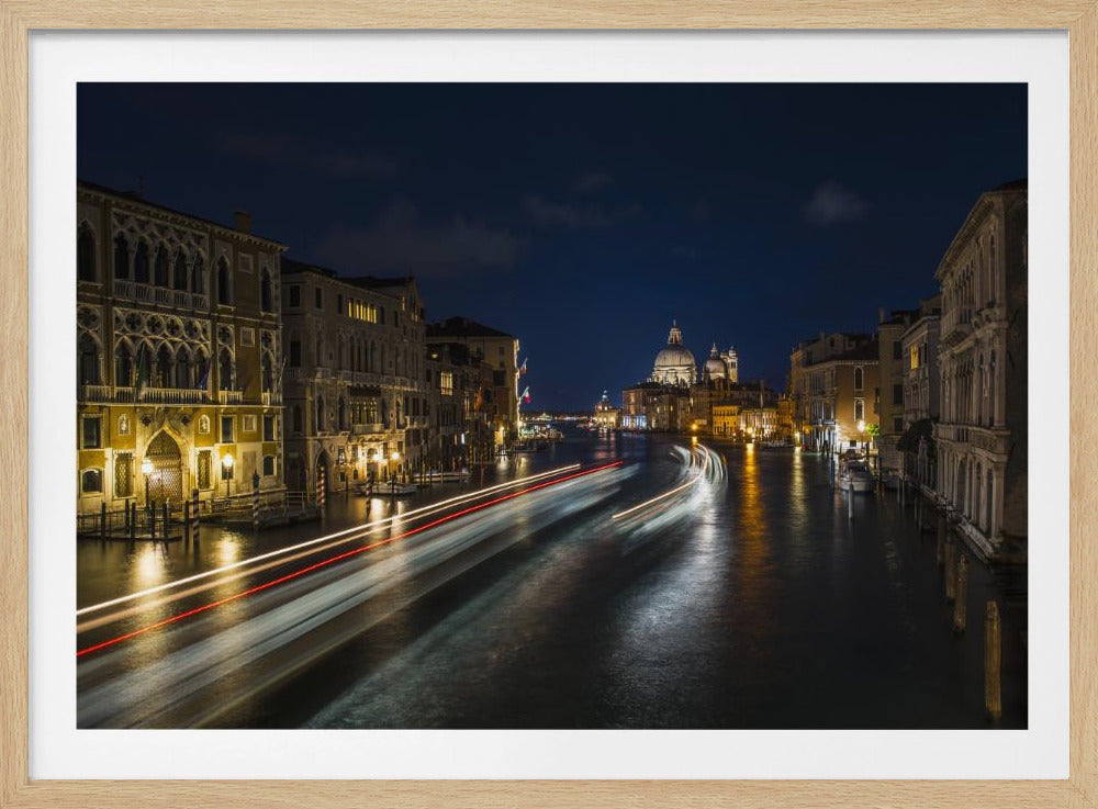 A long-exposure night photograph of the Grand Canal in Venice, Italy. Light trails from moving boats create streaks of red and white on the dark water, which reflects the warm glow of the illuminated historic buildings lining the canal. In the distance, the dome of the Basilica di Santa Maria della Salute stands out against the deep blue night sky. The entire scene is presented within a silver picture frame. Artwork