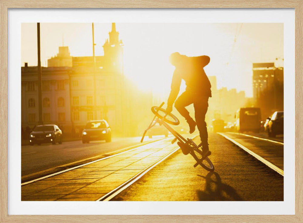 A silhouetted BMX rider performs a trick on a city street during a brilliant golden sunset, with the sun flaring behind them and casting long shadows on the tram tracks. Decor