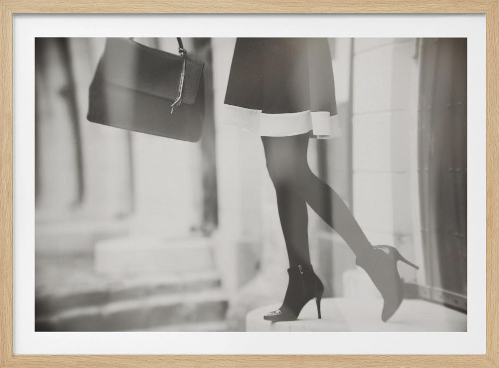 A framed, black and white fashion photograph focusing on the lower half of a woman as she walks. She is wearing a dark skirt with a white hem, black tights, and stylish high-heeled ankle boots, and she is carrying a dark handbag. Print