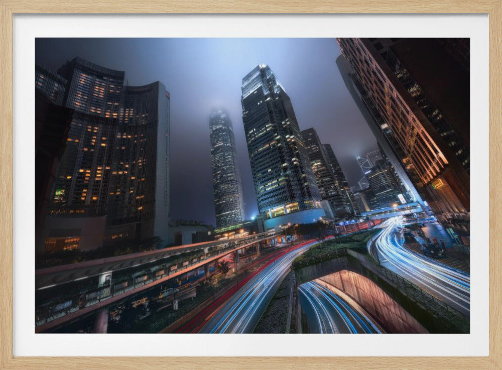 A dynamic, low-angle, long-exposure shot of a modern city at night. Towering skyscrapers with illuminated windows pierce through a foggy, dark blue sky. Below, curving highways are filled with the vibrant red and white light trails of moving traffic. Print