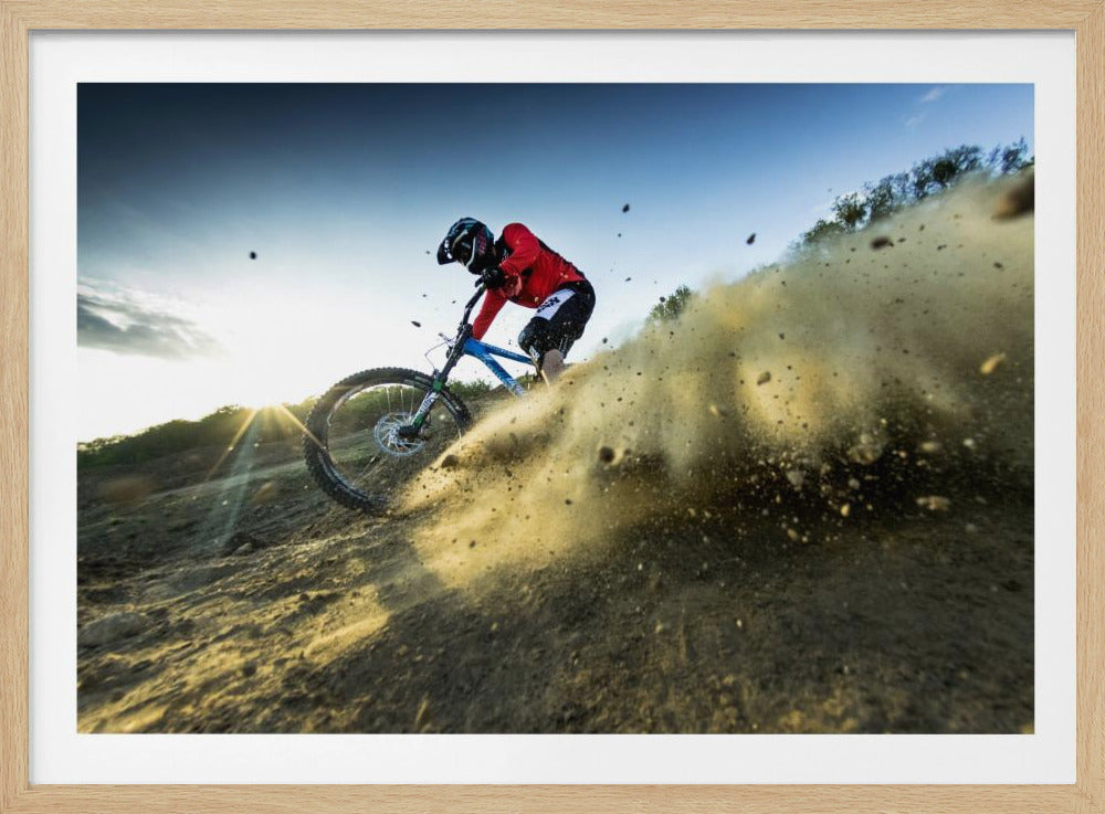 A mountain biker wearing a red jersey and helmet speeds down a steep, dusty trail, kicking up a large cloud of dirt. The sun is setting in the background, creating a lens flare against a clear blue sky. The image is captured from a low angle, emphasizing the action and speed, and is presented within a silver frame. Artwork