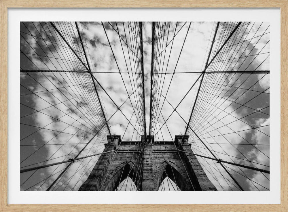 A low-angle, symmetrical black and white photograph looking up at the iconic Brooklyn Bridge. The stone tower with its two Gothic arches is centered, while the complex web of suspension cables radiates outwards and upwards against a dramatic, cloudy sky. The image is presented within a silver frame. Artwork