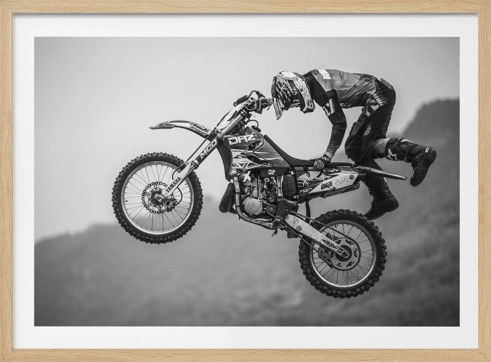 A dynamic black and white photograph of a motocross rider in mid-air, performing a jump. The rider, wearing full protective gear and a helmet, is leaning forward over the handlebars of the dirt bike. The bike is suspended against a blurry, light gray background, emphasizing the action and height of the jump. The image is enclosed in a silver frame. Wall Art