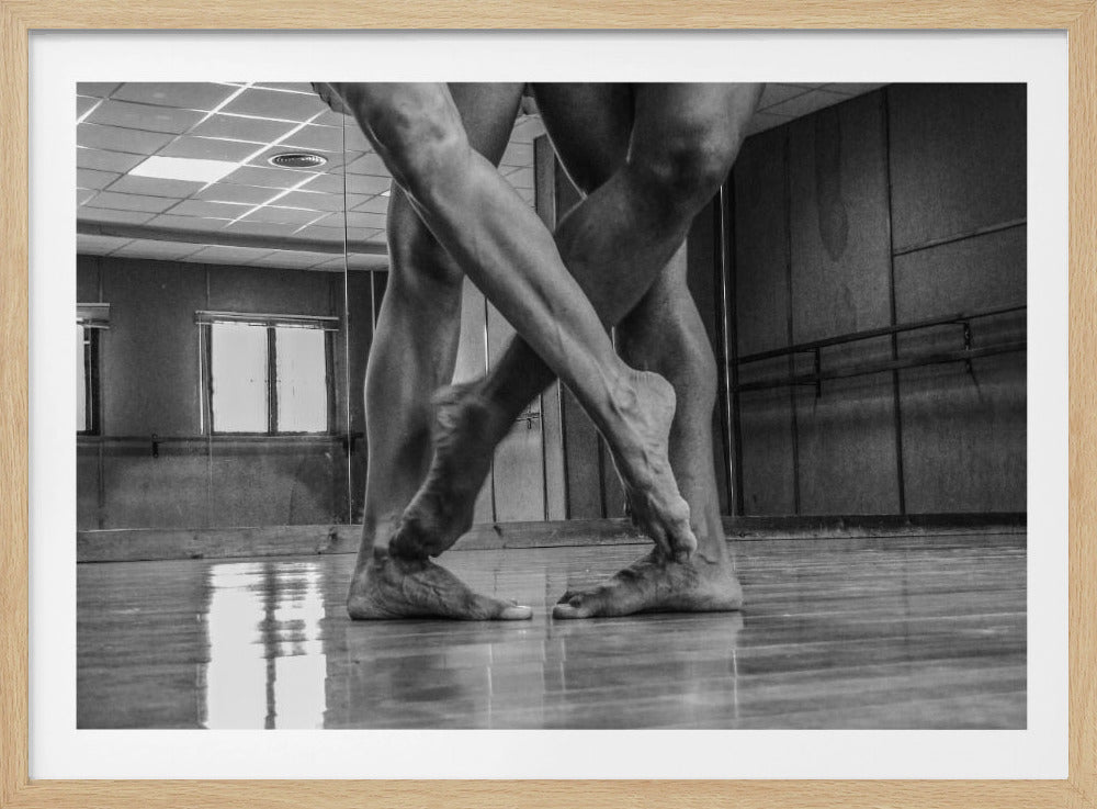 A dramatic low-angle, black and white photograph capturing the muscular legs and bare feet of two dancers intertwined in a dance studio. Their limbs cross in a display of strength and grace on a reflective wooden floor, with the studio's mirrored wall and barre visible in the background. The image is enclosed in a silver-grey frame. Decor