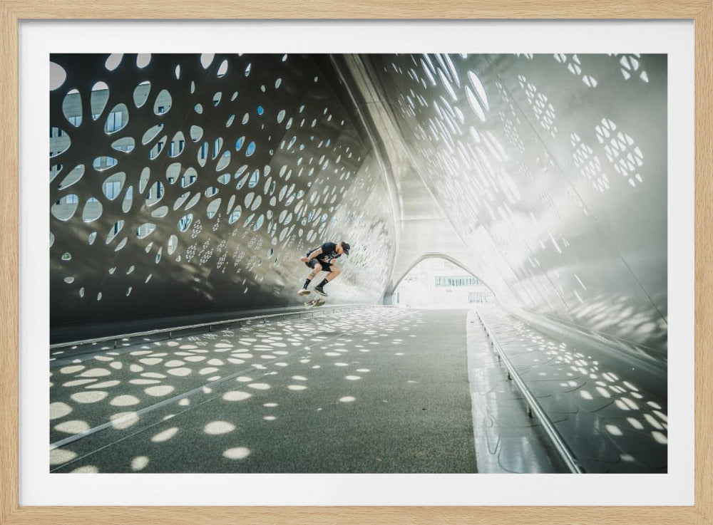 A skateboarder is captured mid-air performing a trick inside a futuristic, metallic tunnel with patterned cutouts that cast dappled light and shadows across the scene, all enclosed within a silver frame. Artwork
