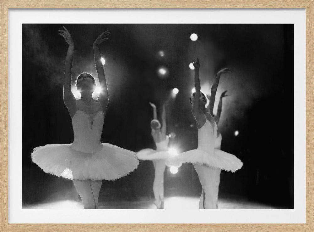 A dramatic black and white photograph of a group of ballerinas in white tutus performing on a dark, hazy stage. The ballerina in the foreground is backlit by a spotlight, with her arms gracefully raised above her head. Other dancers are visible in the background, similarly posed under stage lights. The entire image is presented within a silver, textured frame. Decor