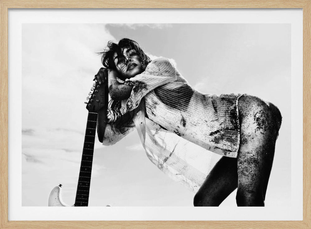 A dramatic, low-angle black and white photograph of a woman with disheveled hair, wearing a sand-covered shirt, leaning exhaustedly against the neck of an electric guitar against a bright sky. Wall Art