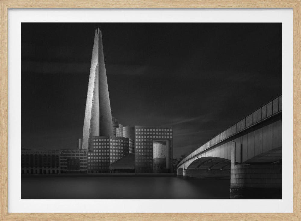 A dramatic black and white photograph of the London skyline featuring The Shard skyscraper and London Bridge. The image is shot at dusk or night, with The Shard brightly lit against a dark, moody sky. The River Thames in the foreground is calm and reflective, captured with a long exposure. Artwork